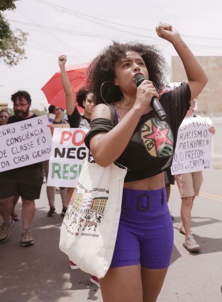 Jaque Medeiros fala ao microfone em ato de rua, com o punho erguido.